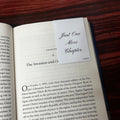 Open book with a magnetic bookmark labeled 'Just One More Chapter' on a wooden surface
